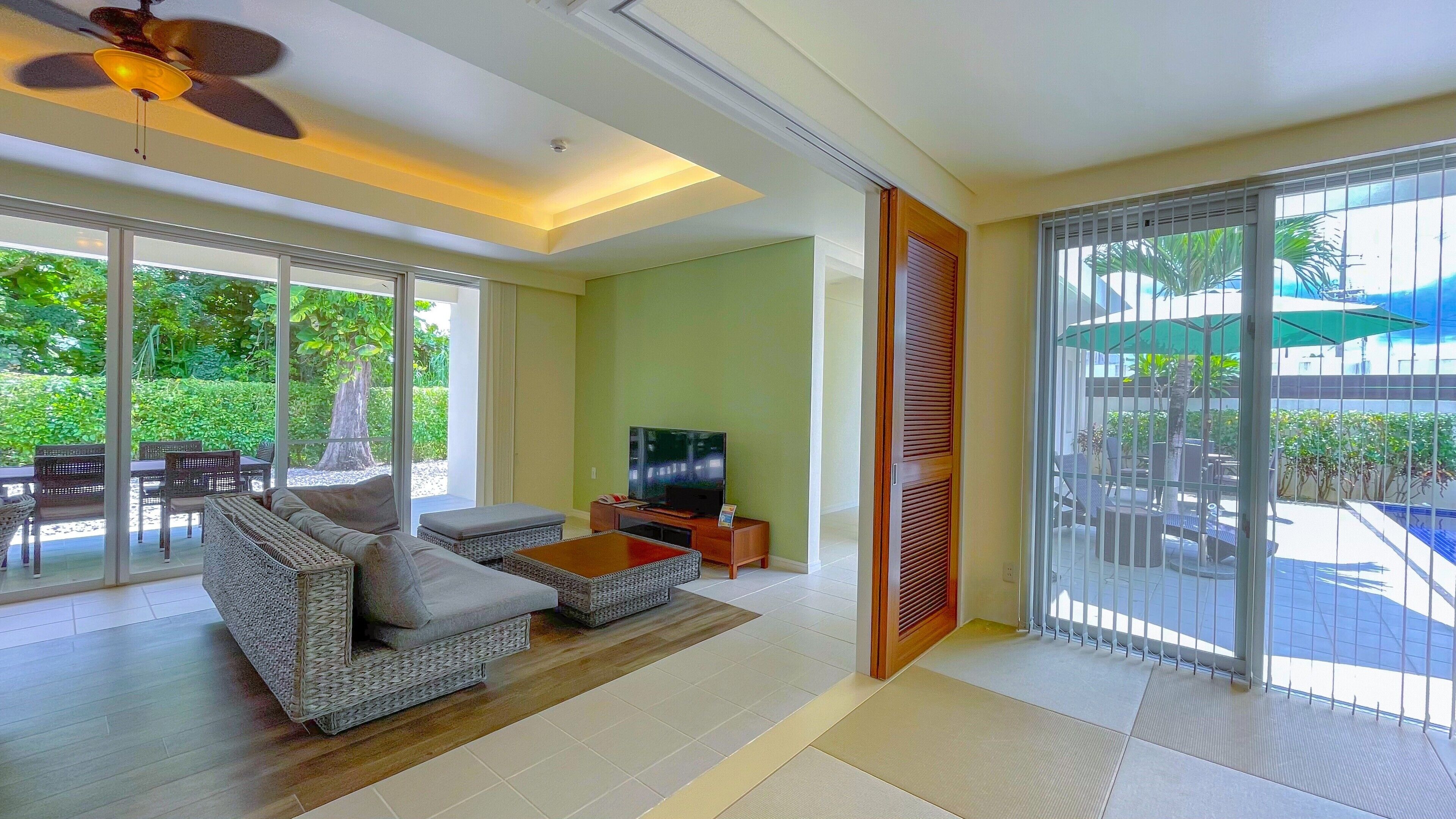 A spacious living room with a sofa, coffee table, and TV, large sliding glass doors leading to a balcony with outdoor furniture.