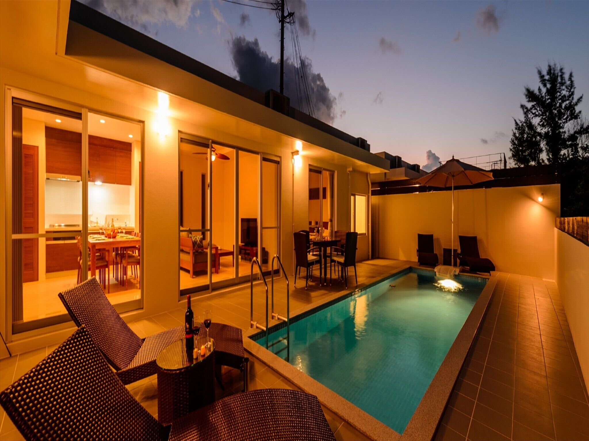 A modern outdoor pool area with a dining setup and a kitchen visible through glass doors.
