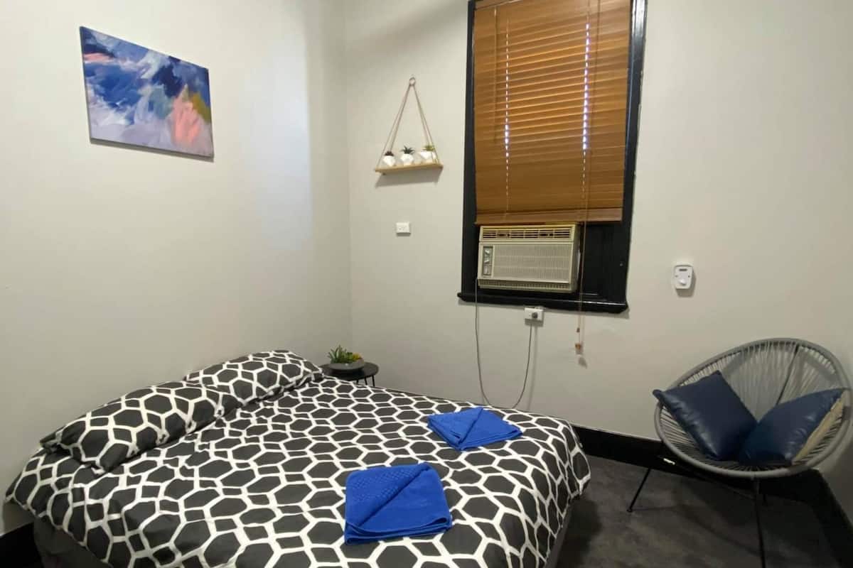 A bedroom with a bed, a ceiling fan, a window with blinds, a chair, and a wall-mounted air conditioner.
