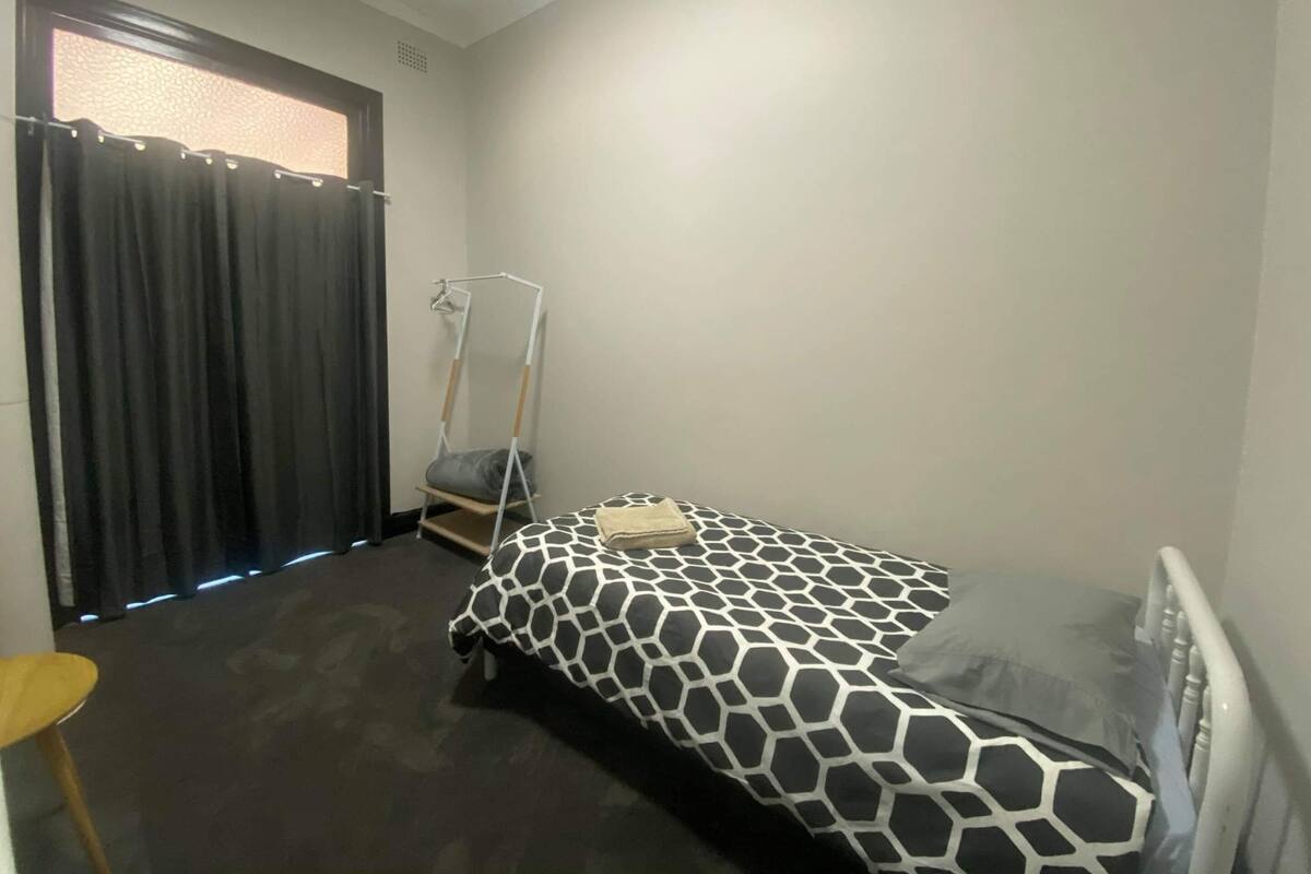 A small, single-bed room with a black and white patterned bedspread, a standing desk with a chair, a window with curtains, and a plain wall.
