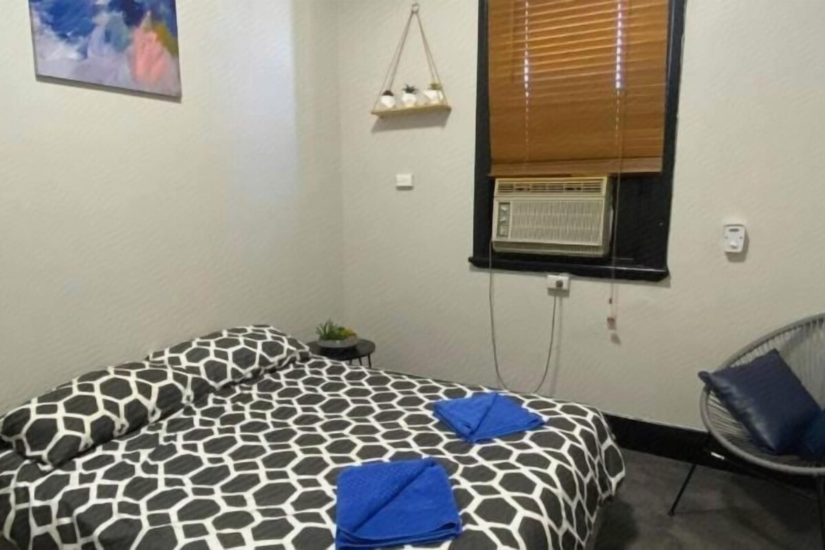 A bedroom with a bed, a chair, a window with blinds, and a wall-mounted air conditioner.