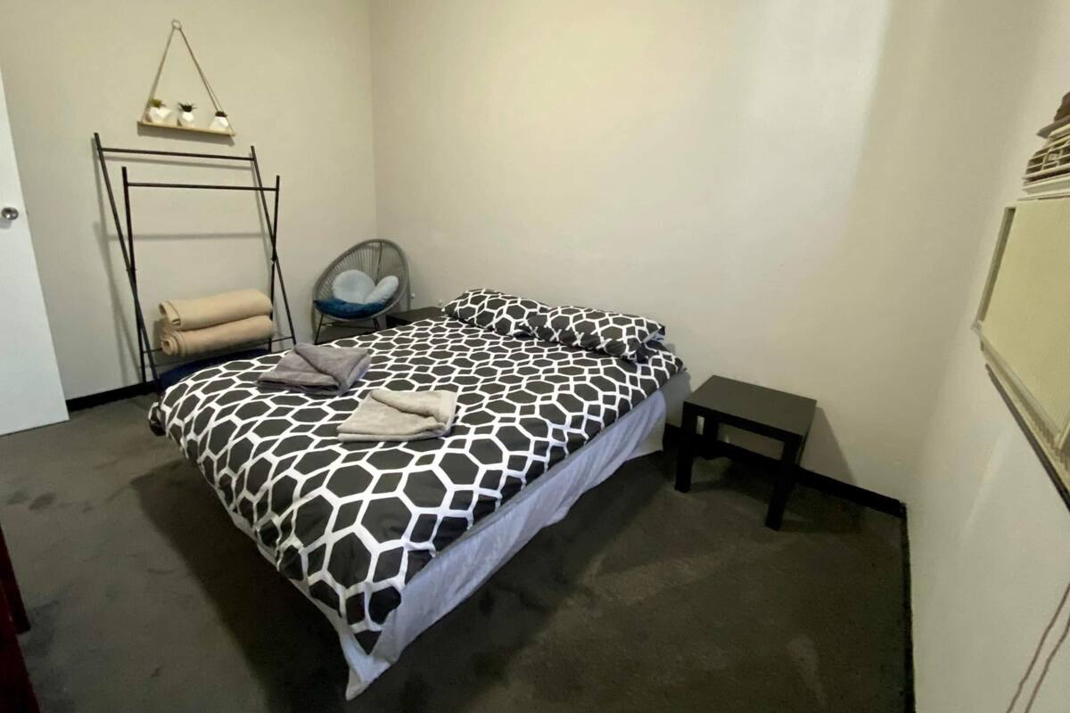 A bedroom with a bed, a small table, a chair, a shelf with items, and a radiator.