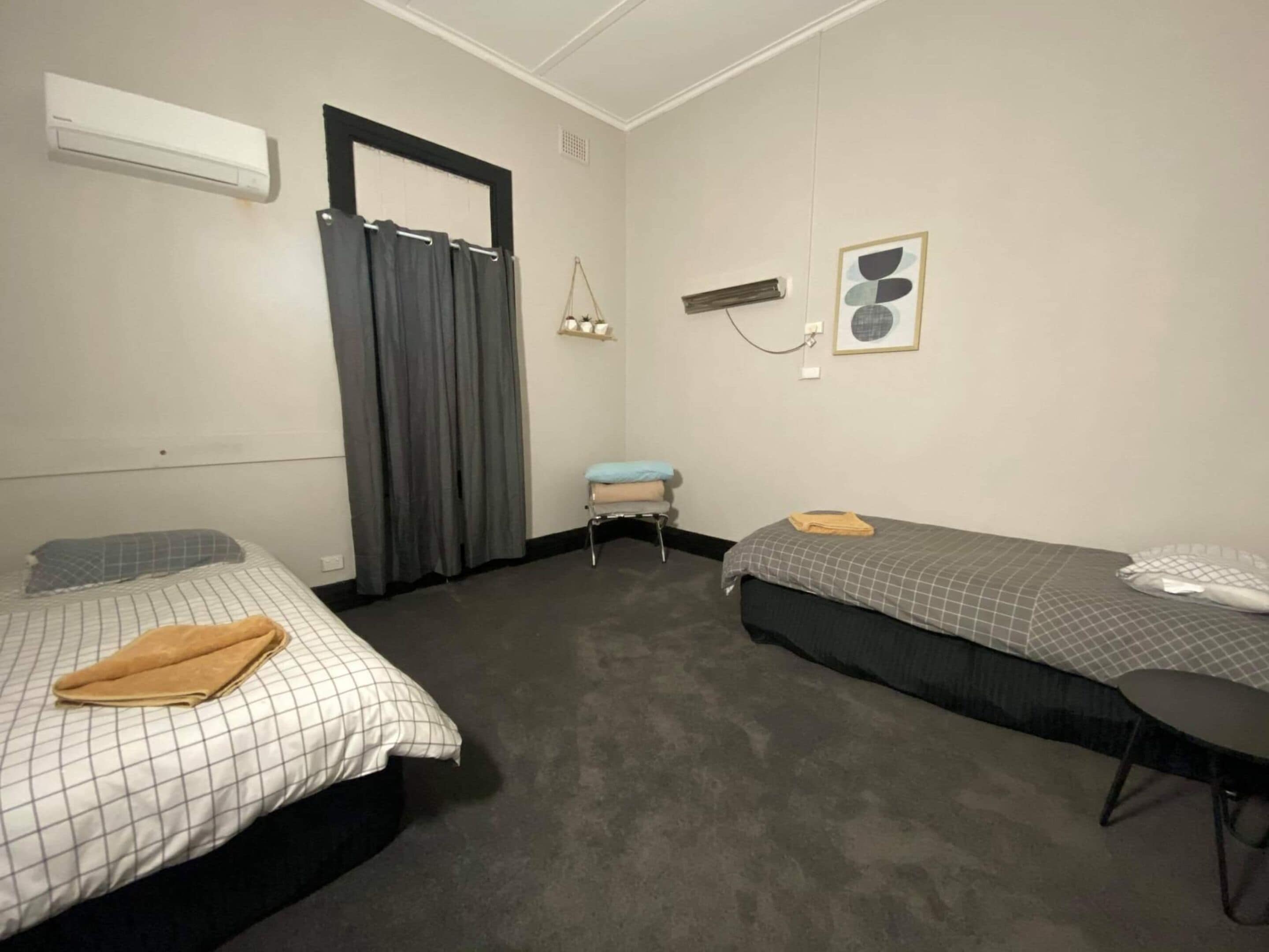 Une petite chambre propre, équipée de deux lits simples, d’un bureau et d’une chaise. On y trouve une fenêtre avec des rideaux, un climatiseur fixé au mur et une petite étagère avec des articles de toilette.