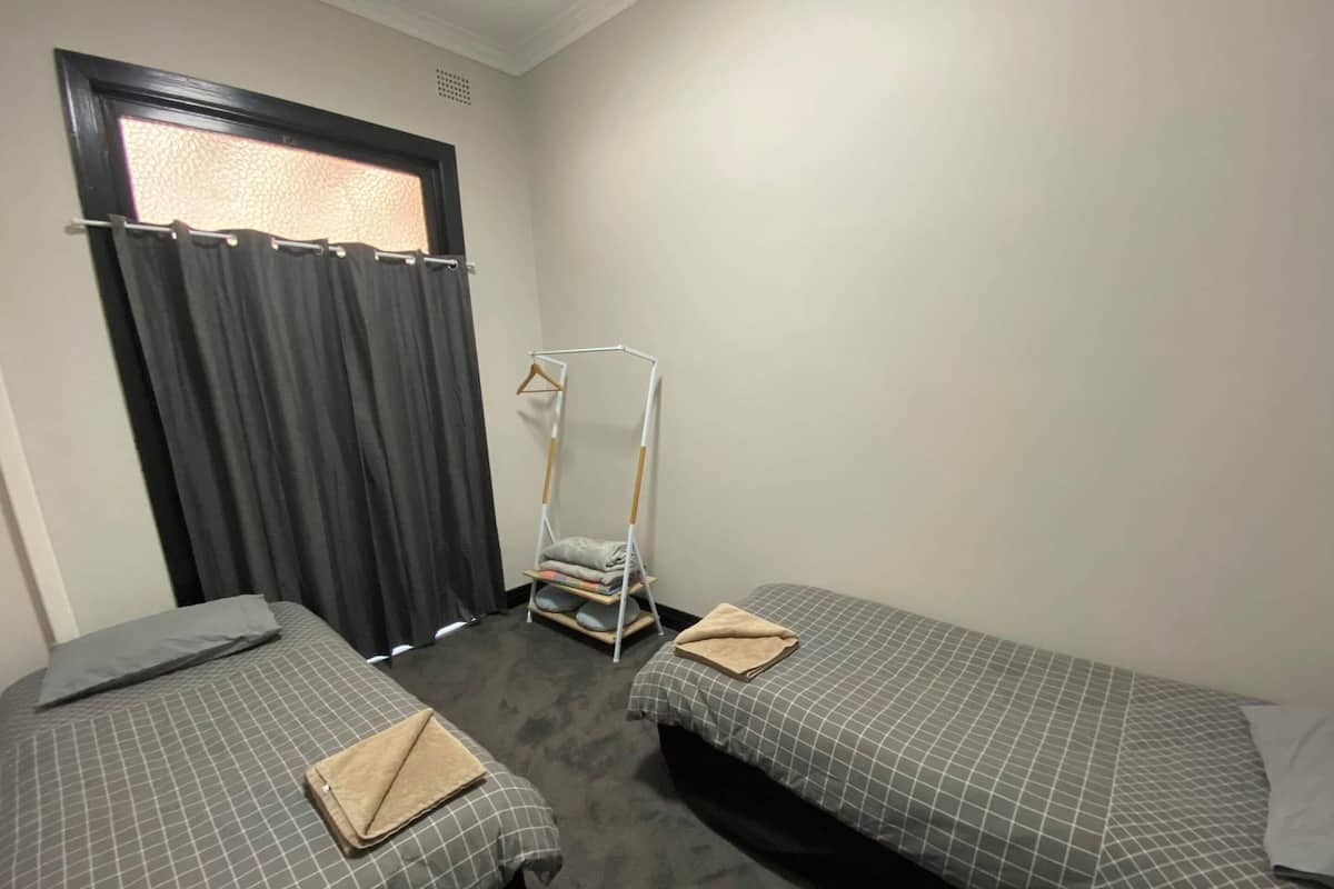 A room with two single beds, a window with curtains, a standing shelf unit, and a radiator.