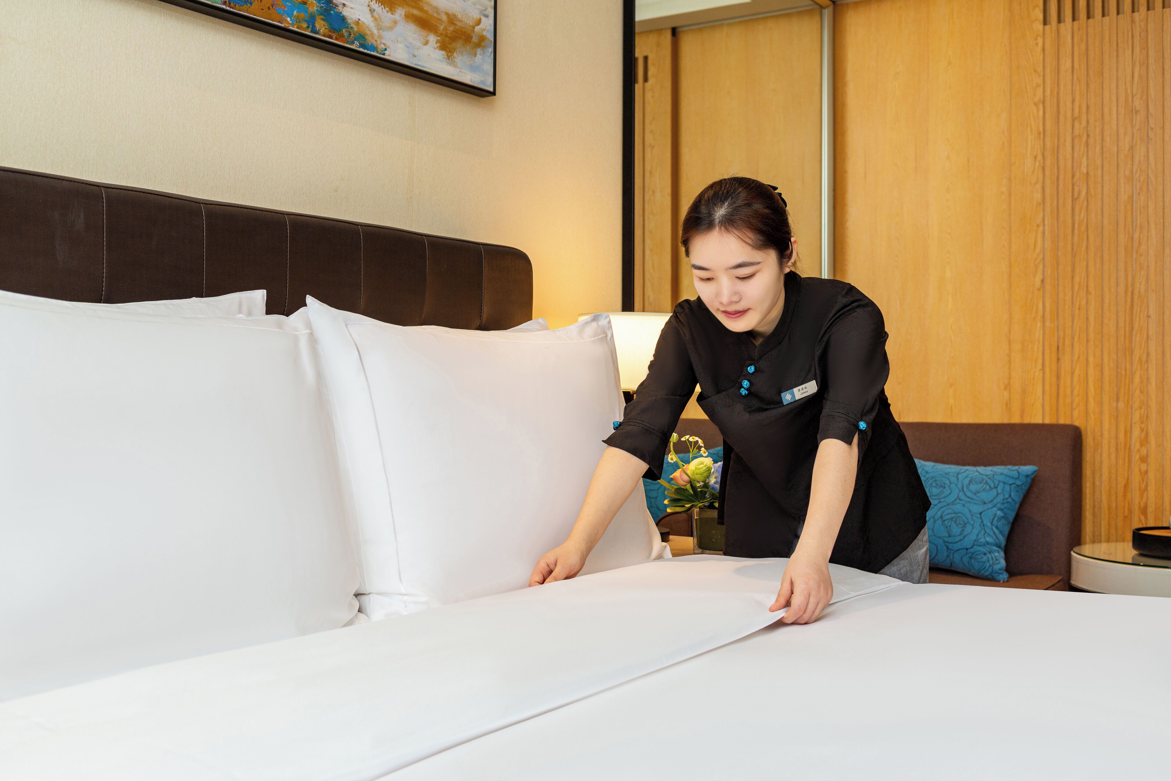 A person adjusting a bed in a hotel room with a wooden headboard, a blue patterned pillow, and a framed picture on the wall.
