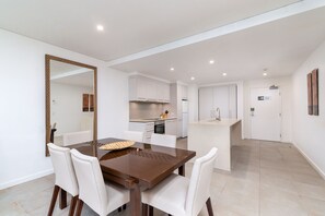 Una cocina moderna con armarios blancos, una isla central y un comedor con un espejo grande y sillas blancas.