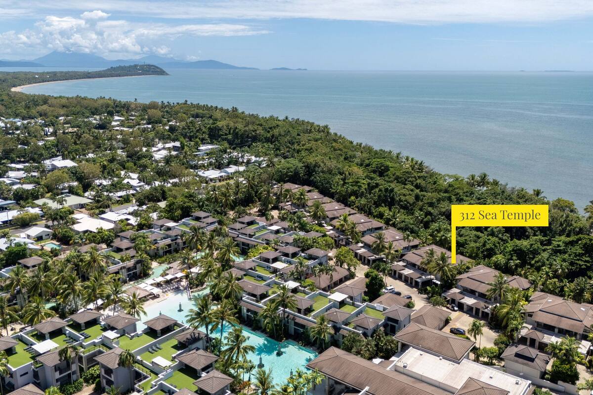 Vista aérea de un complejo turístico con varias cabañas, una piscina central y vistas al mar.