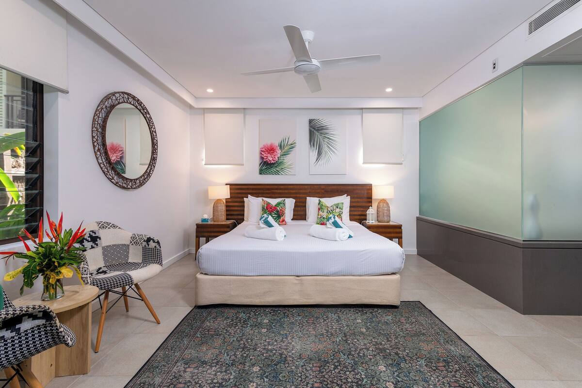 Un dormitorio moderno con una cama grande, dos mesitas de noche con lámparas, un ventilador de techo, un espejo decorativo y vistas al exterior.
