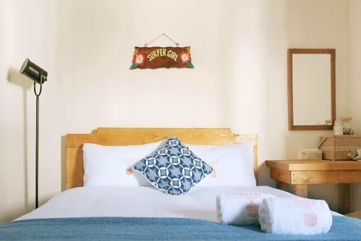 A neatly made bed with a blue blanket, a patterned pillow, and a sign above reading "SURFER GIRL".