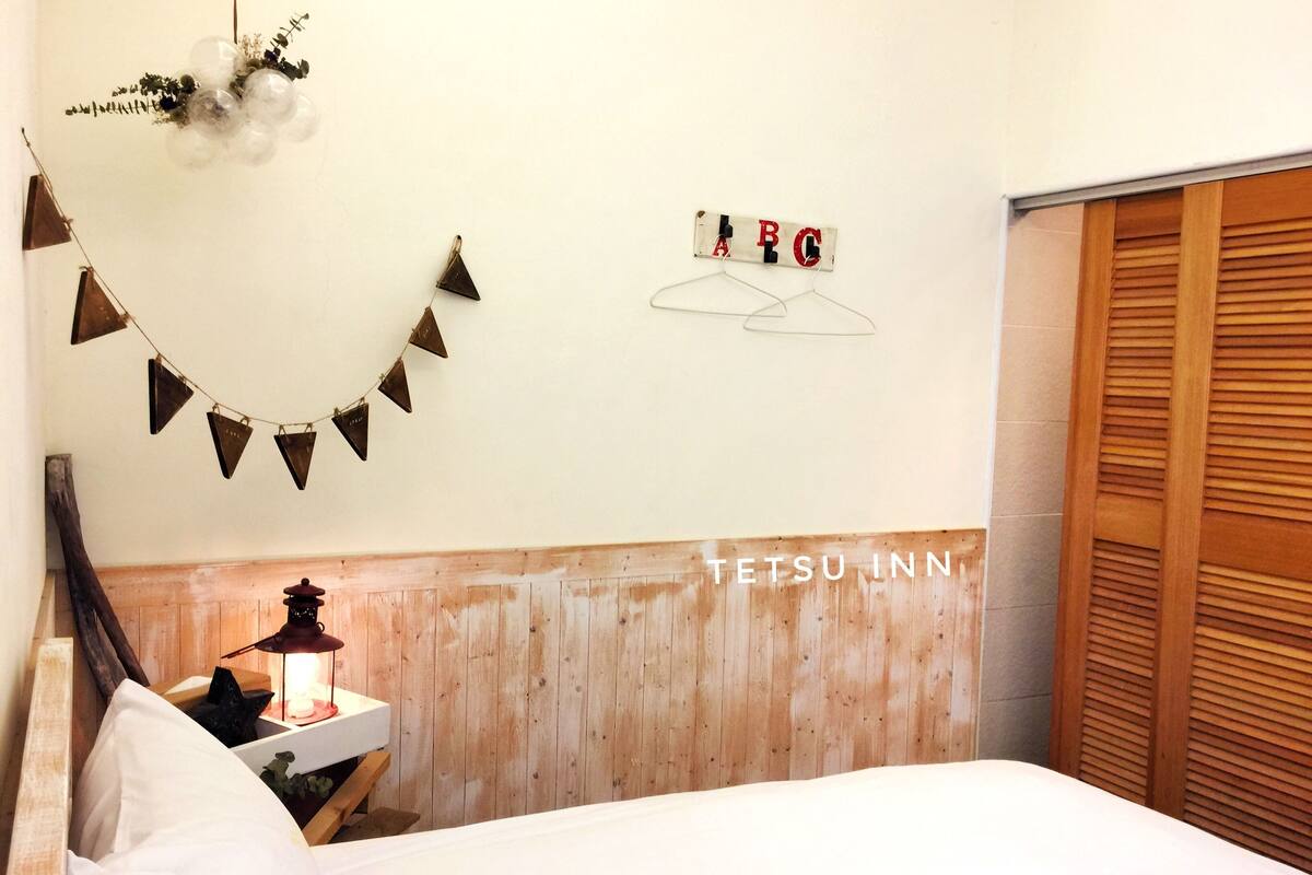 A bedroom with a wooden headboard, a bedside table with a lantern, a wall-mounted letter 'A' and 'B' sign, and a hanging string of triangular decorations.