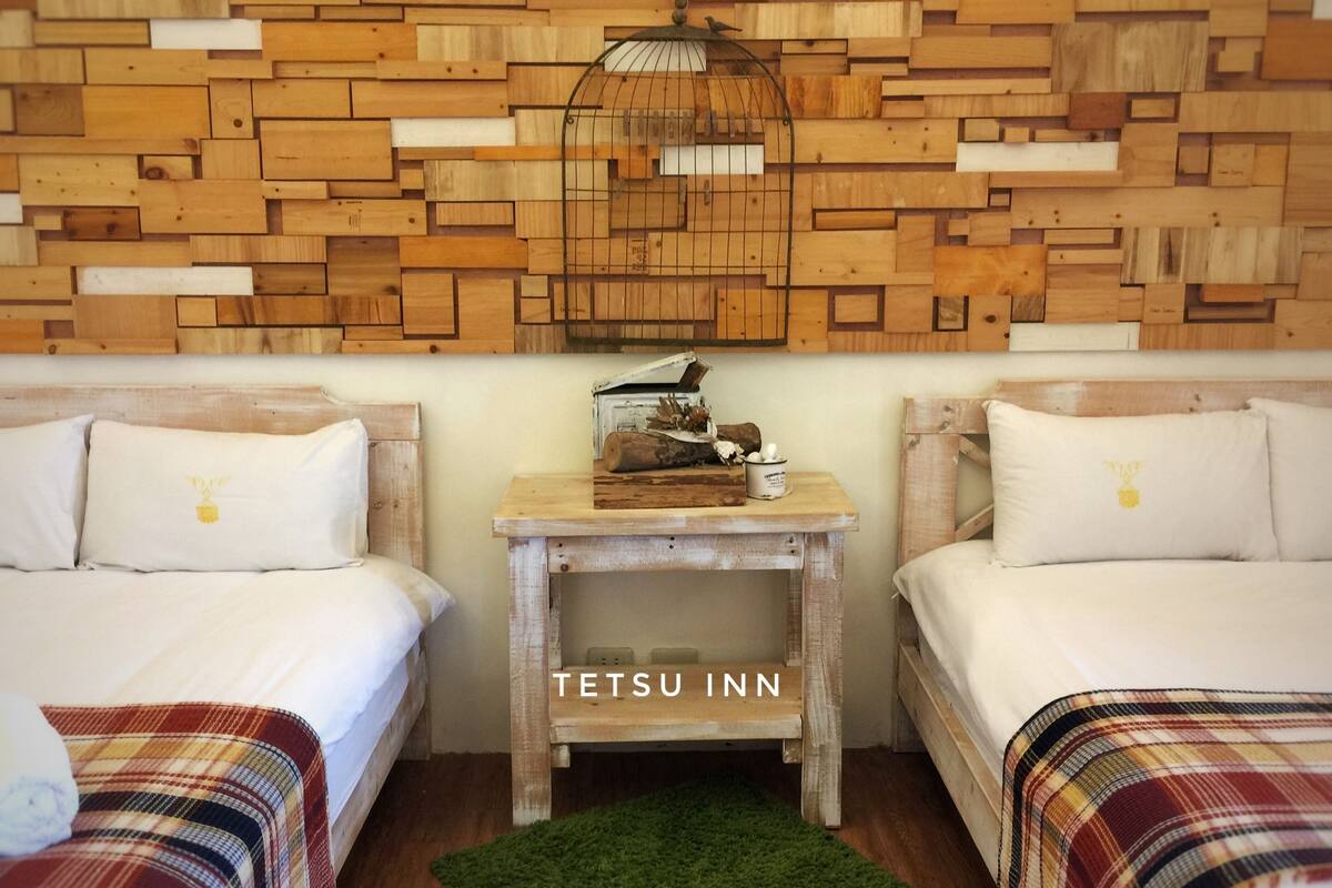 A room with two beds, a wooden wall, a small table with a birdcage, and a green rug.