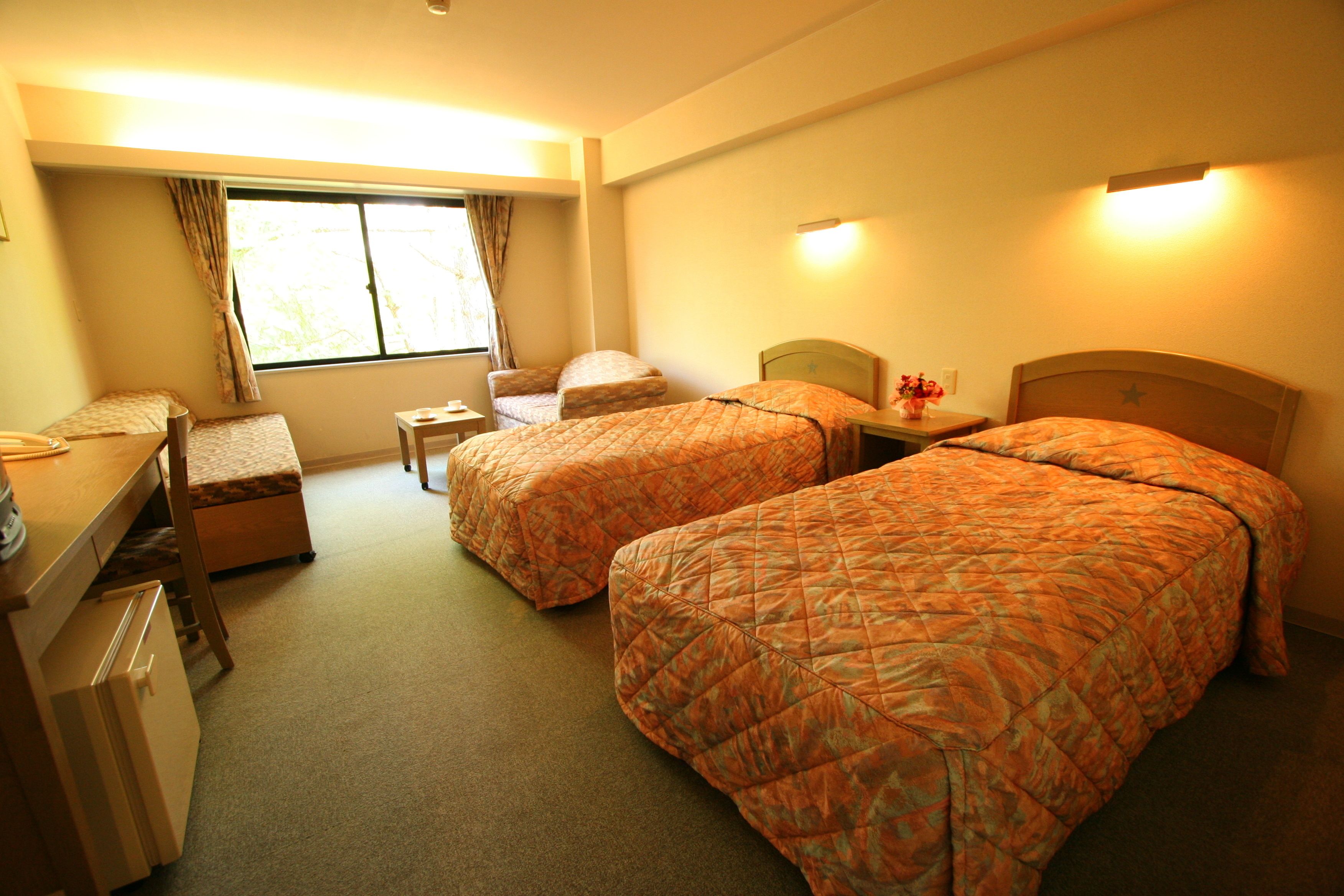 Ein Hotelzimmer mit zwei Betten, einem Schreibtisch, einem Stuhl, einem kleinen Tisch und einem Fenster mit Vorhängen.