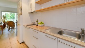 A modern kitchen with white cabinets, a stainless steel sink, wooden shelves, and a dining area with a table and chairs. - Seascape Holidays at Portsea (Port Douglas)