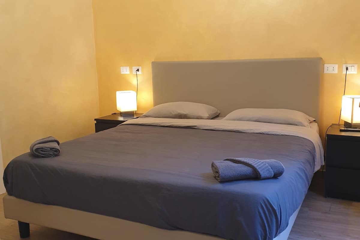 A neatly made bed with a grey and white duvet, two pillows, and a folded towel on the left side. There are two bedside tables with lamps, and the walls are painted yellow.