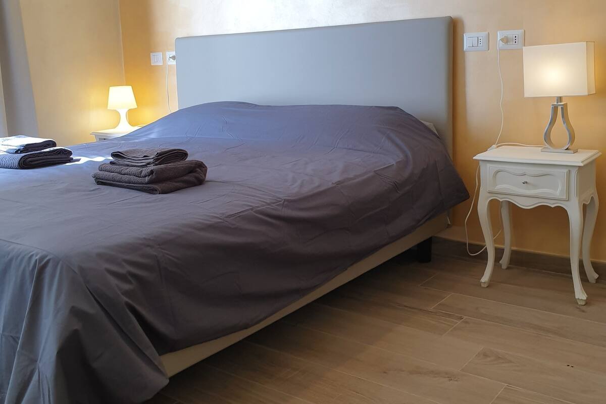 A neatly made bed with a grey bedspread and folded towels, a white nightstand with a lamp, and a wall socket.