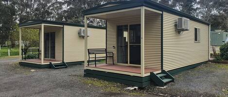 Two small, single-story cabins with a covered porch and a bench. Each cabin has a sliding glass door and an air conditioning unit mounted on the exterior wall.