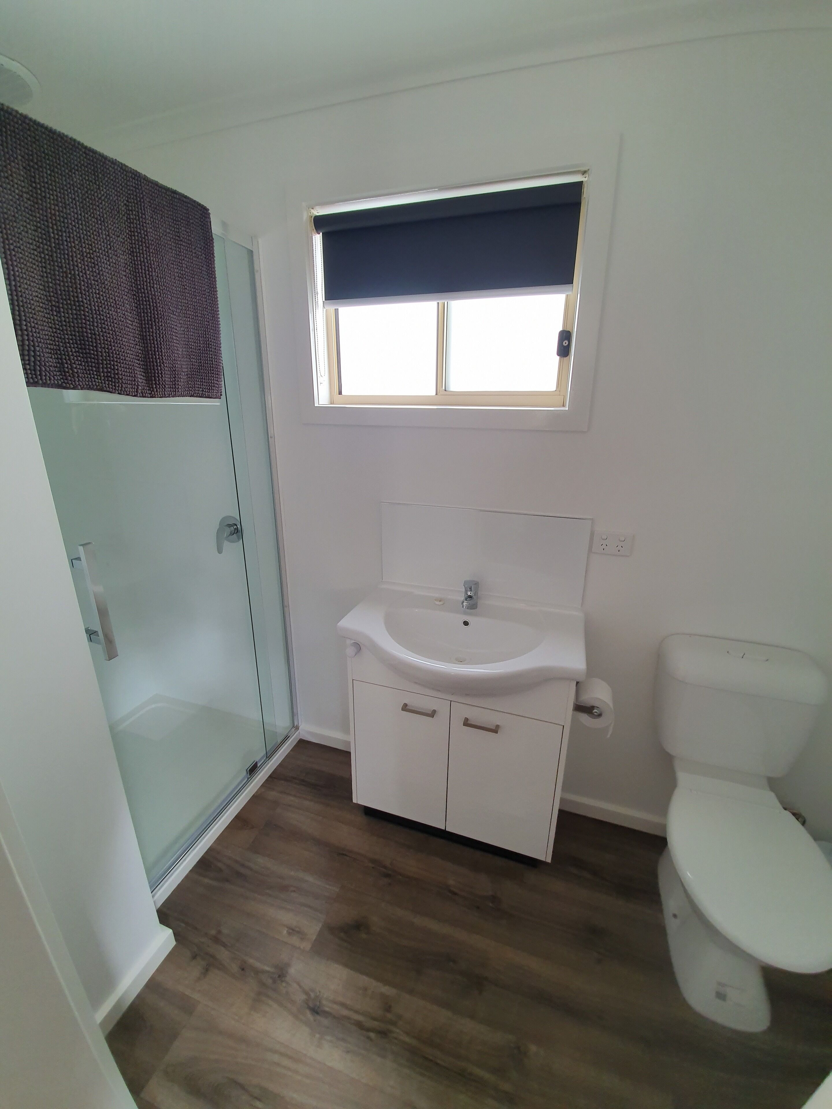 A bathroom with a glass shower enclosure, a white sink with a window above, and a toilet.