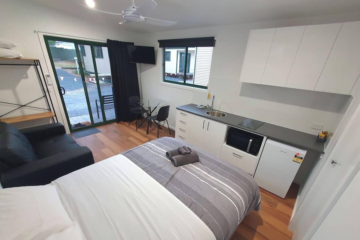 A compact hotel room with a bed, a small kitchenette, a dining area, and a sliding glass door to the outside.
