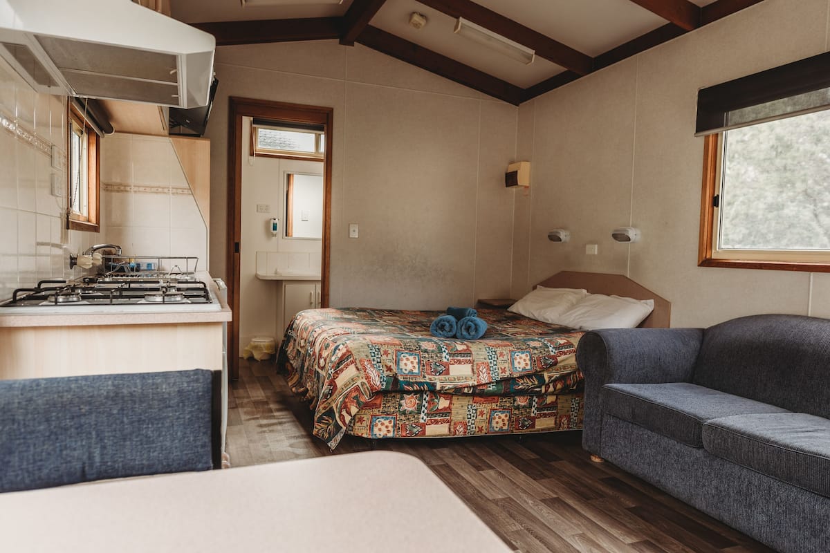 A compact room with a bed, a sofa, a small kitchen area with a stove, and a window providing natural light.
