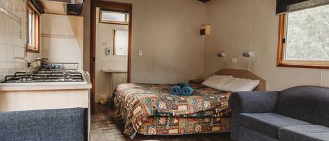Habitación compacta con cama, sofá, pequeña cocina con estufa y ventana que proporciona luz natural.