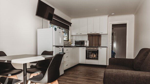 A modern kitchen with white cabinets, a microwave, and a built-in oven. There is a dining table with chairs and a sofa in the living area. - G'day Parks Ararat (Ararat)