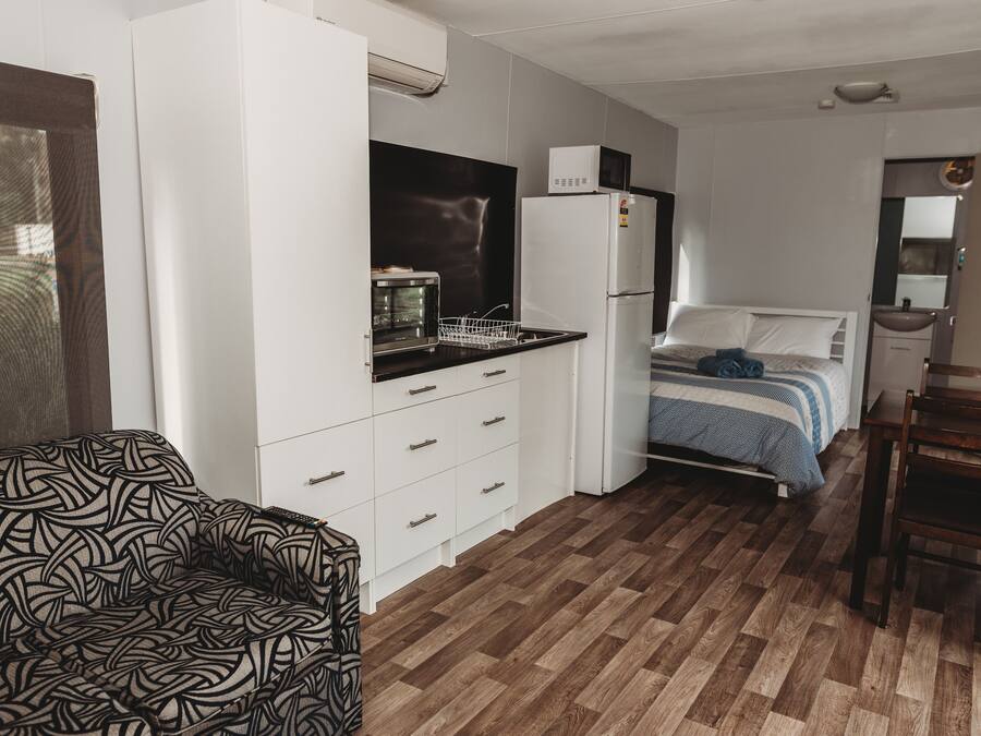 A compact room with a kitchenette, a bed with a blue and white striped bedspread, a wooden floor, and a patterned chair.