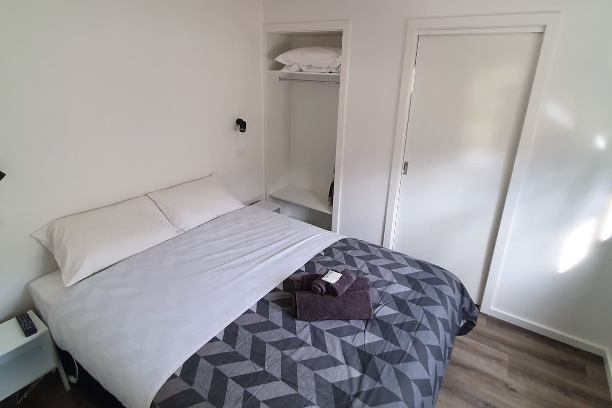 A neatly made bed with a patterned bedspread, a white pillow, and a folded towel on the bed. There is a white nightstand with a remote control beside the bed. A closet with a hanging rod and a door is visible in the background.