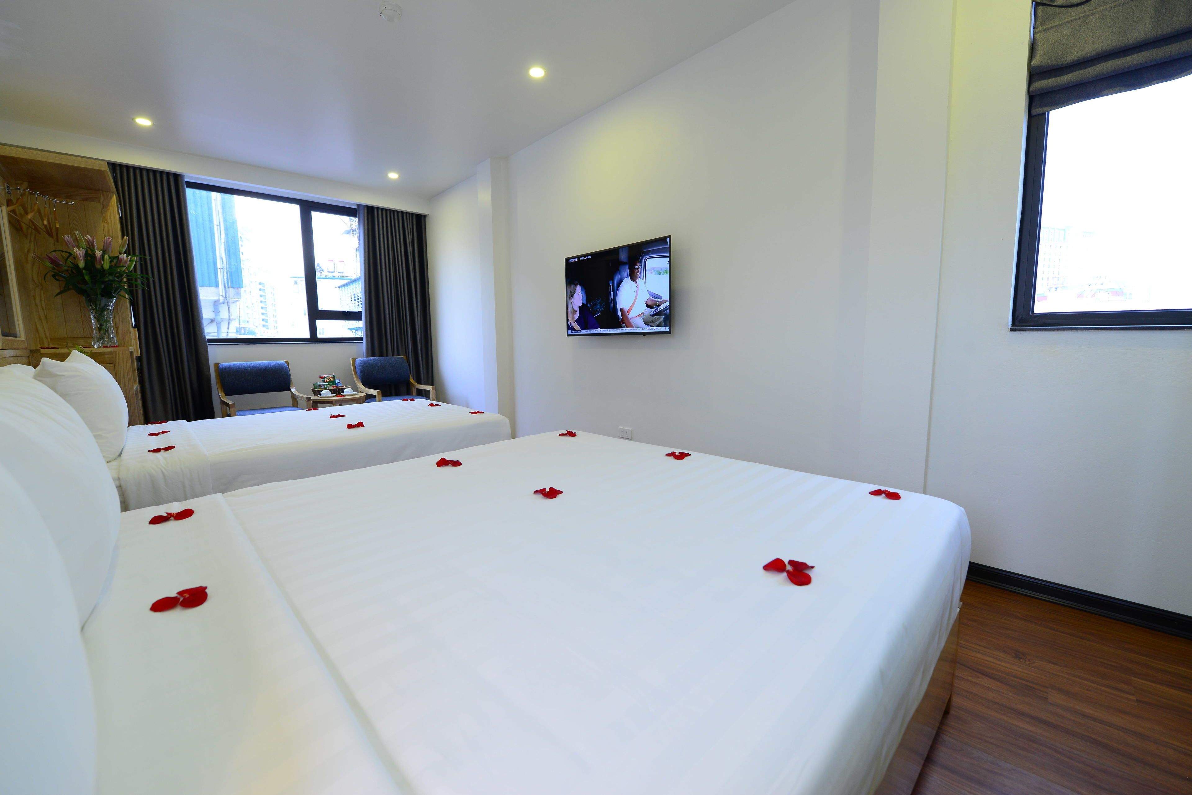 A hotel room with two beds, each adorned with red rose petals. A television is mounted on the wall, and a window provides a view of the cityscape.
