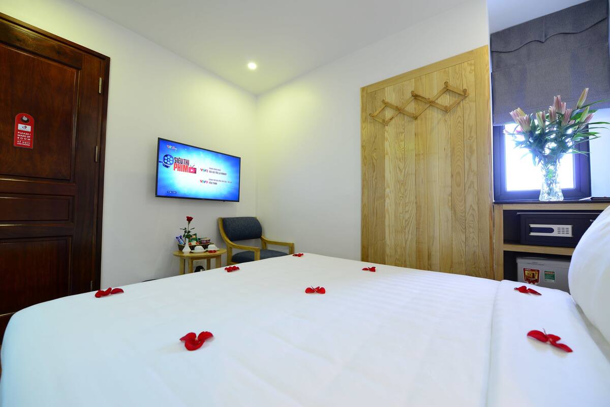 Ein Hotelzimmer mit Bett, Fernseher, Holzschrank, kleinem Tisch mit einer Vase und einem Fenster mit Blumen.