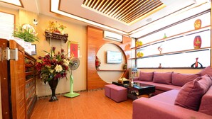 A modern interior with a wooden reception desk, a large potted plant, a pink sofa, and a television mounted on a wooden panel.