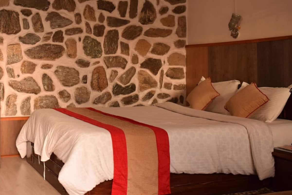Quarto com parede de pedra, cama com roupa de cama branca e detalhe em vermelho, e cabeceira de madeira.
