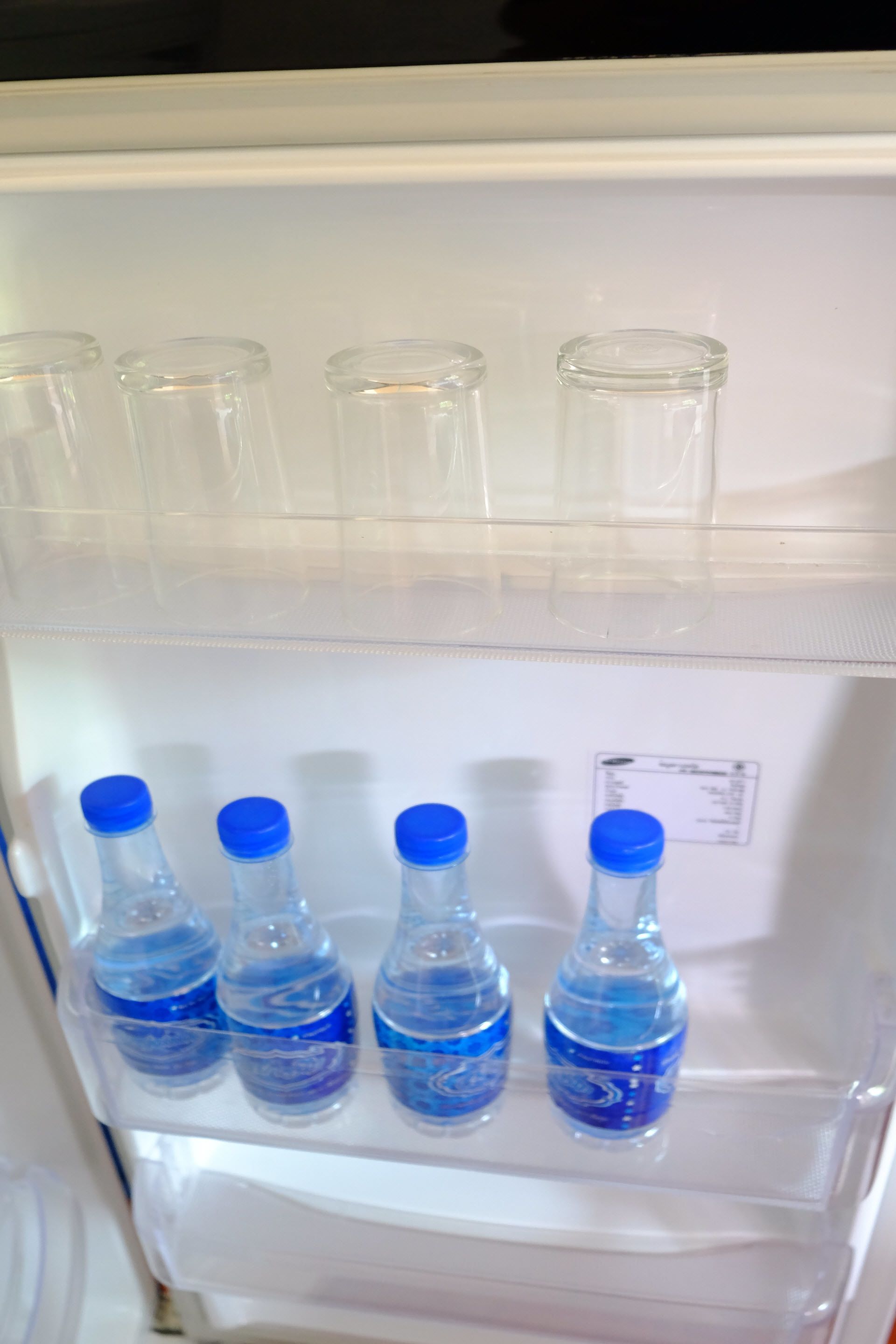 Un refrigerador con dos estantes; el estante superior tiene botellas vacías y el inferior, cinco botellas con tapas azules.