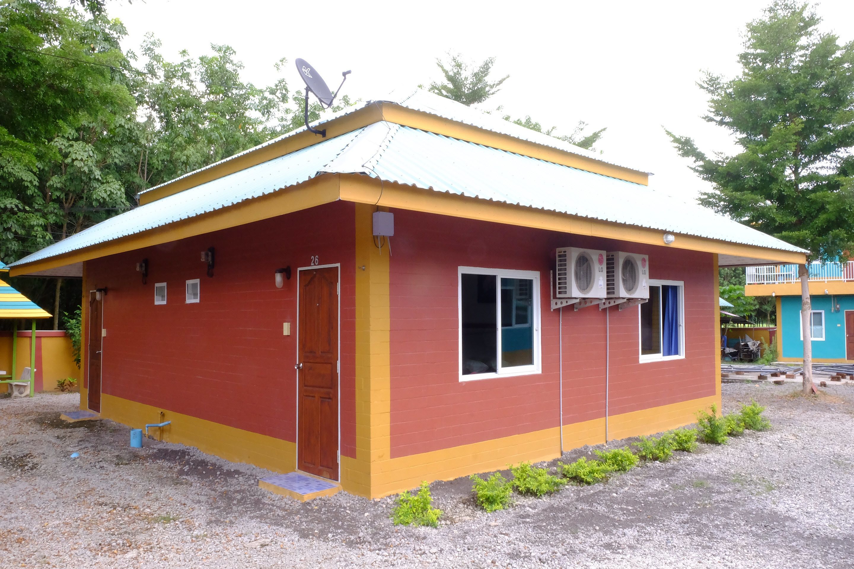 Um edifício pequeno, térreo, com fachada vermelha e amarela, antena parabólica no telhado e terreno de cascalho.