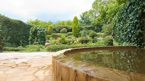 Une zone dallée de pierres, avec une fontaine circulaire au centre, entourée d’une végétation luxuriante et de différentes espèces de plantes.