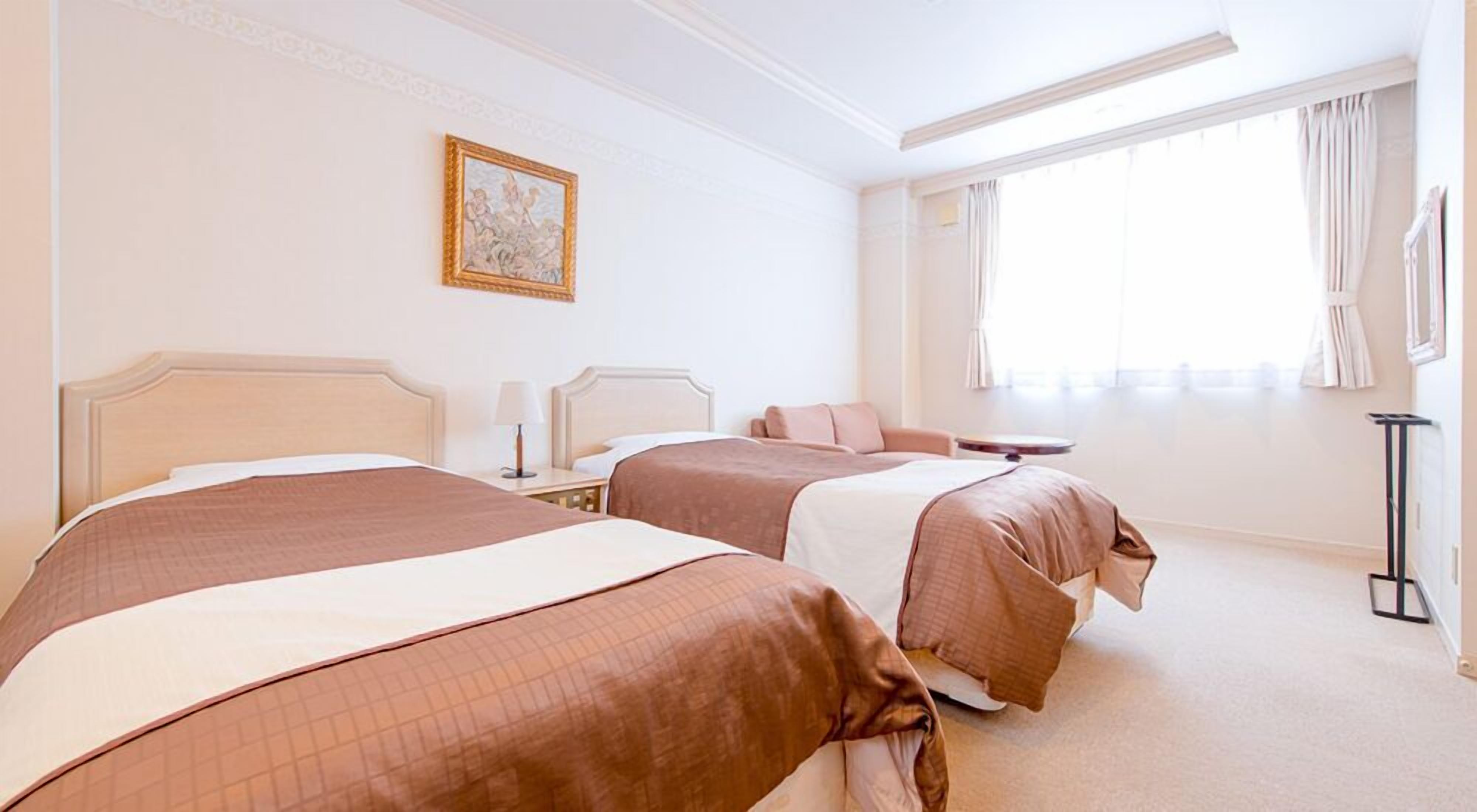 Ein Hotelzimmer mit zwei Betten, einem Nachttisch mit Lampe, einem Bild an der Wand und einem Fenster mit Vorhängen.