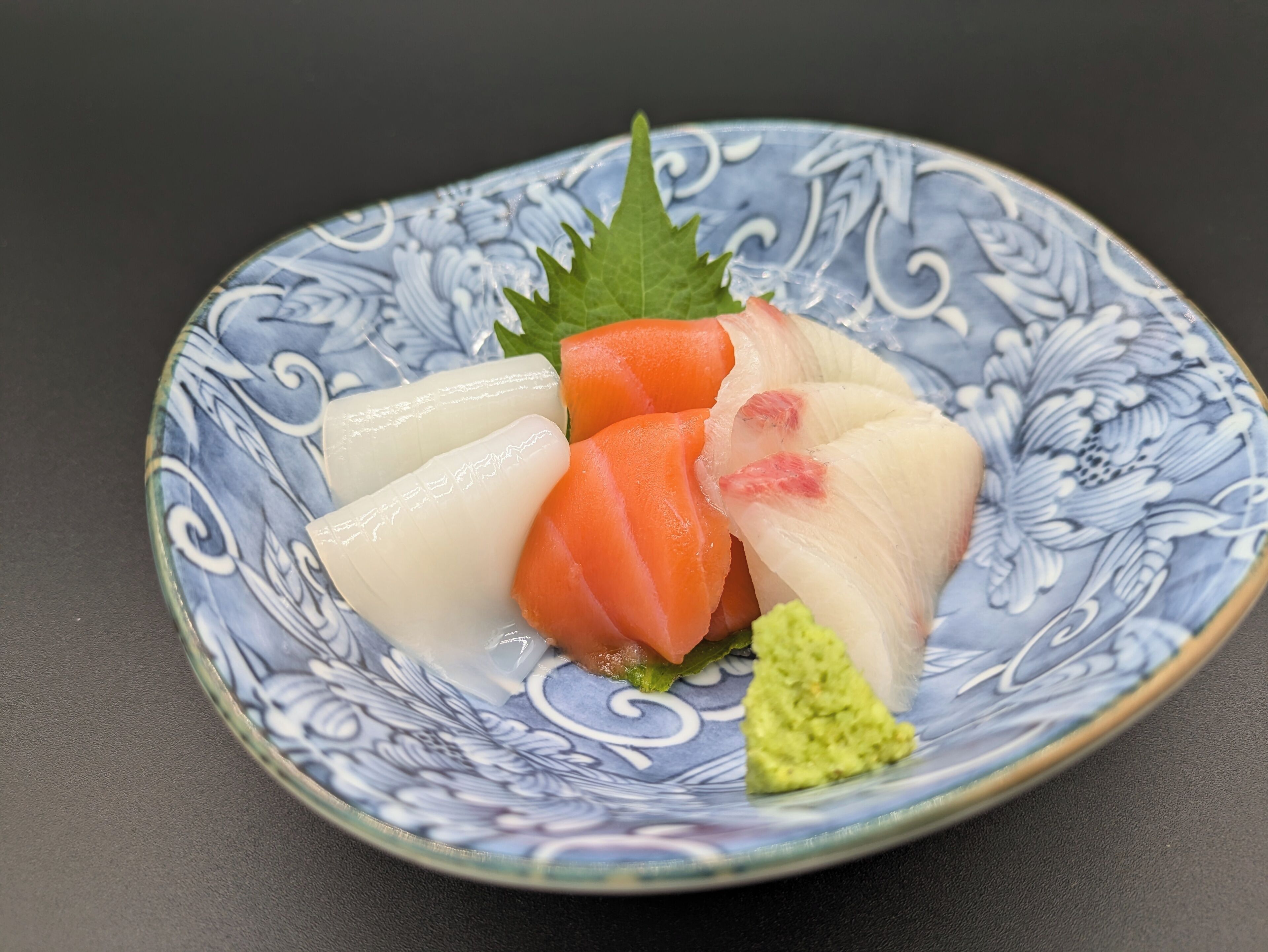 Ein Teller mit Sashimi, darunter weißes Fischfilet, orangefarbener Fisch und grüner Avocado, auf einem dekorativen blau-weißen Teller.