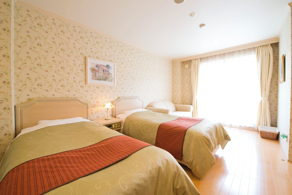 Habitación de hotel con dos camas, papel tapiz floral, un cuadro en la pared y una ventana con cortinas.
