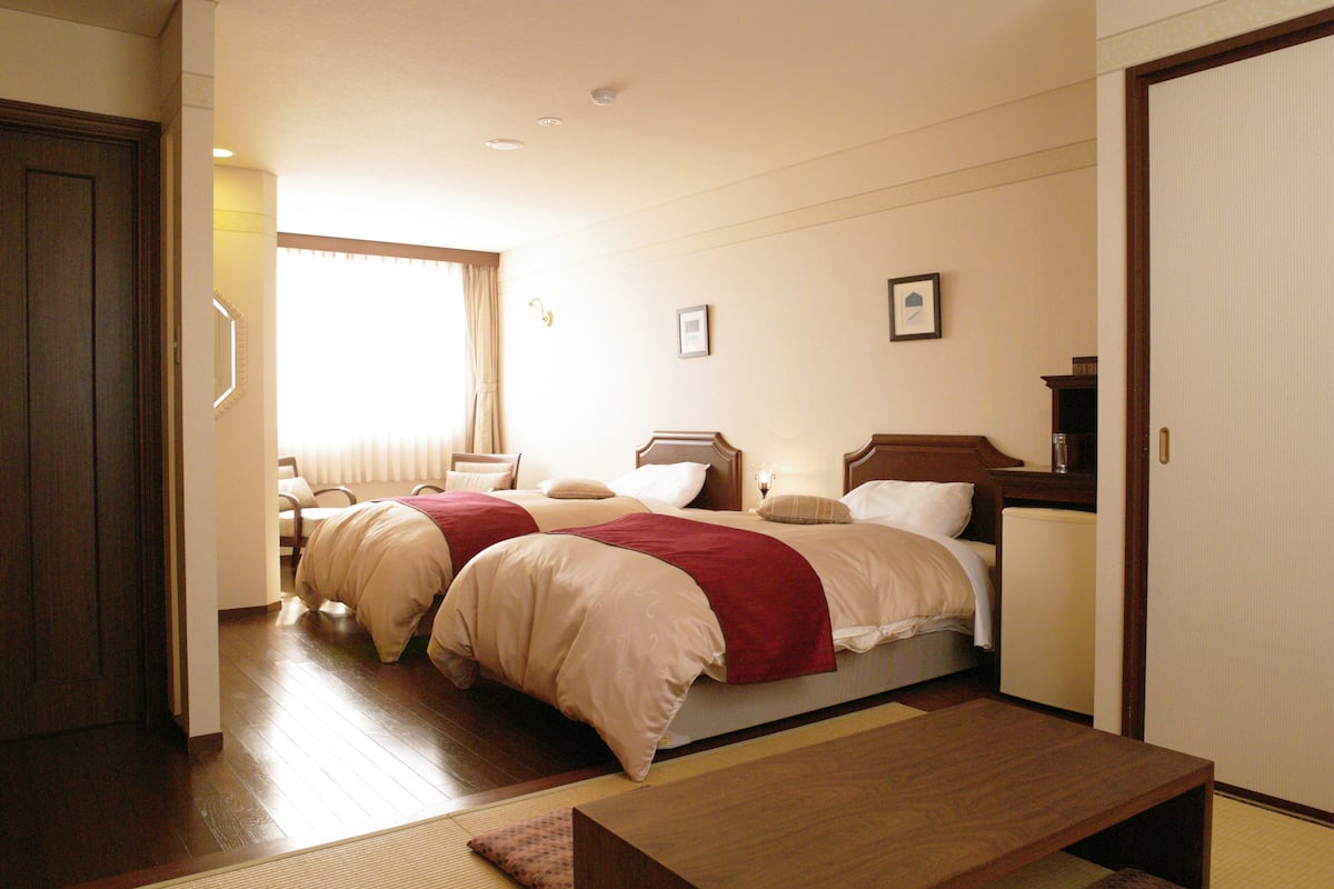 Habitación de hotel con dos camas, un banco de madera, una mesita y un ventanal con cortinas.
