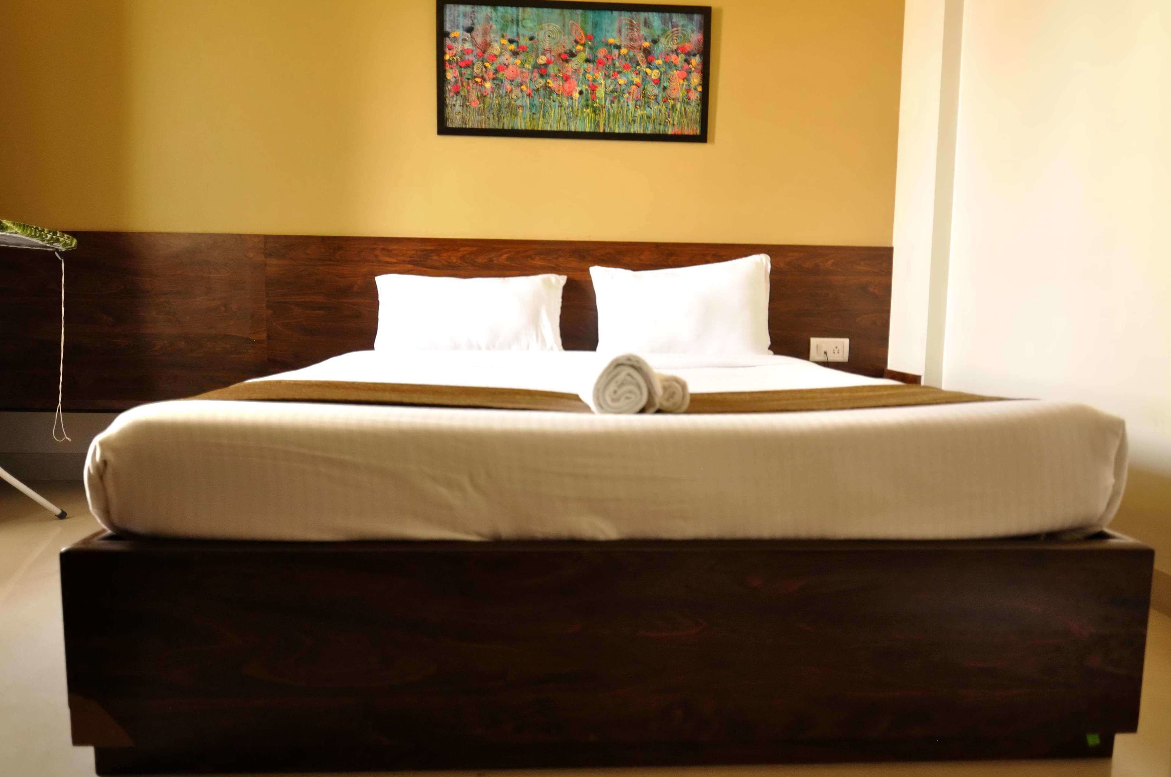 A neatly made bed with white linens and pillows, a wooden headboard, and a painting of flowers on the wall.