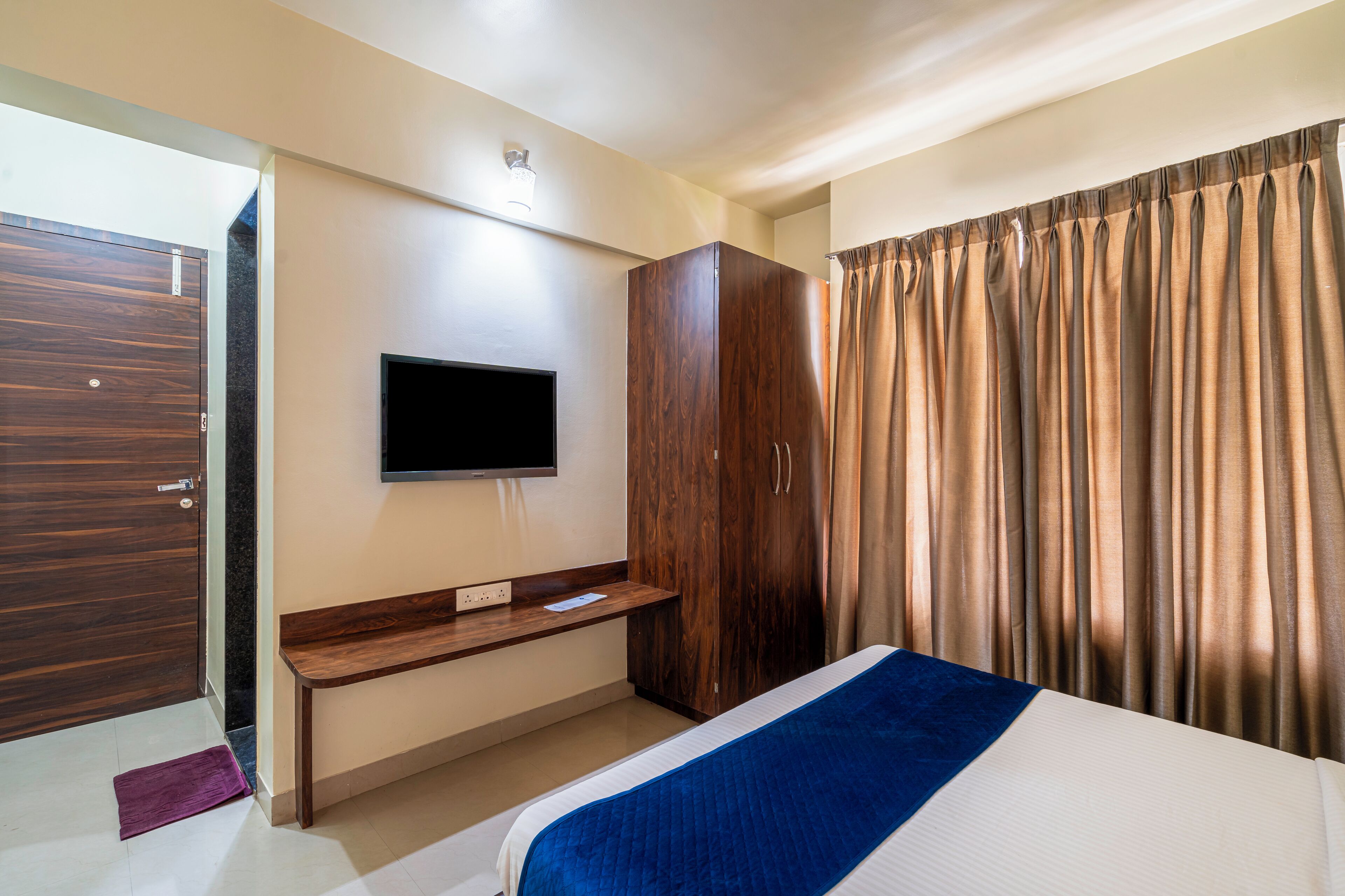 A hotel room with a bed, a TV mounted on the wall, a wooden wardrobe, and a curtain.