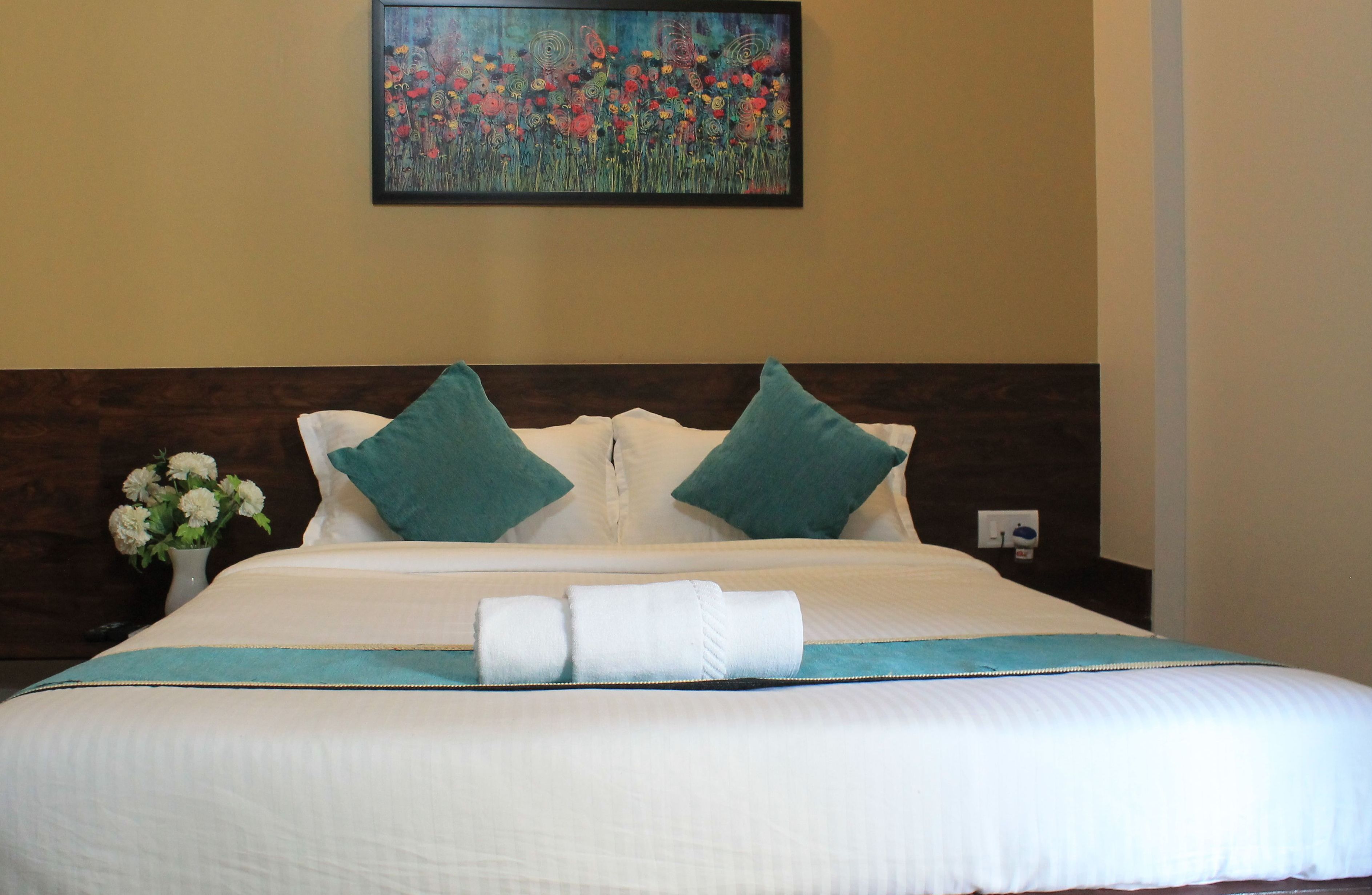 A neatly made bed with white bedding and teal pillows, a vase of flowers on the nightstand, and a painting of flowers on the wall.