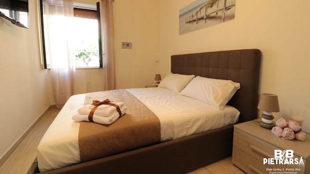 A neatly made bed with a brown headboard and white linens, a bedside table with a lamp and flowers, and a window with sheer curtains. - B&B Pietrarsa (Portici)