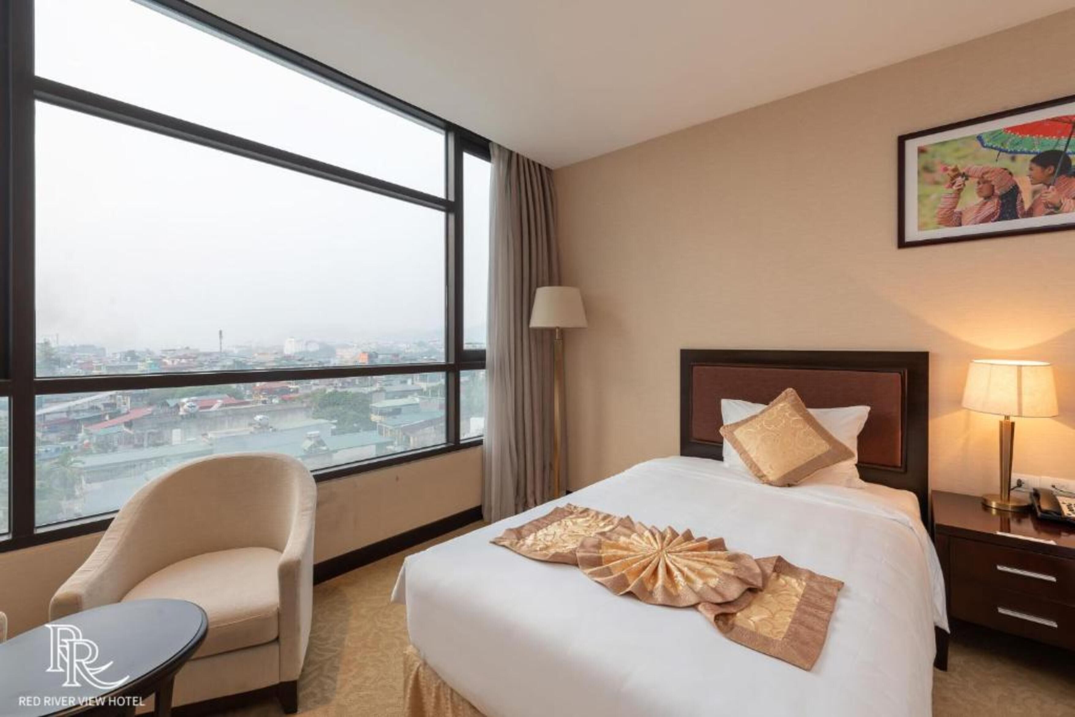 A hotel room with a large window, a bed with a decorative headboard, a bedside table with a lamp, and a chair.