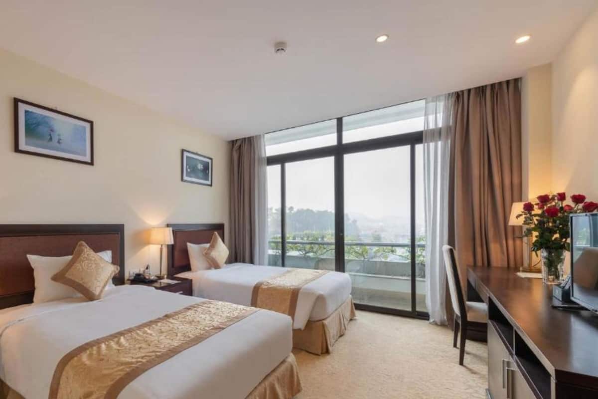 Ein Hotelzimmer mit zwei Betten, einem Schreibtisch, einem Stuhl und einem großen Fenster mit Blick auf die Stadt.