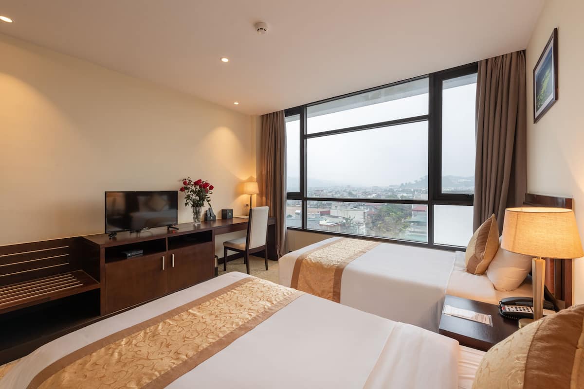 Ein Hotelzimmer mit zwei Betten, einem Schreibtisch mit Fernseher, einem Sessel, einer Lampe und Blick auf die Stadt.