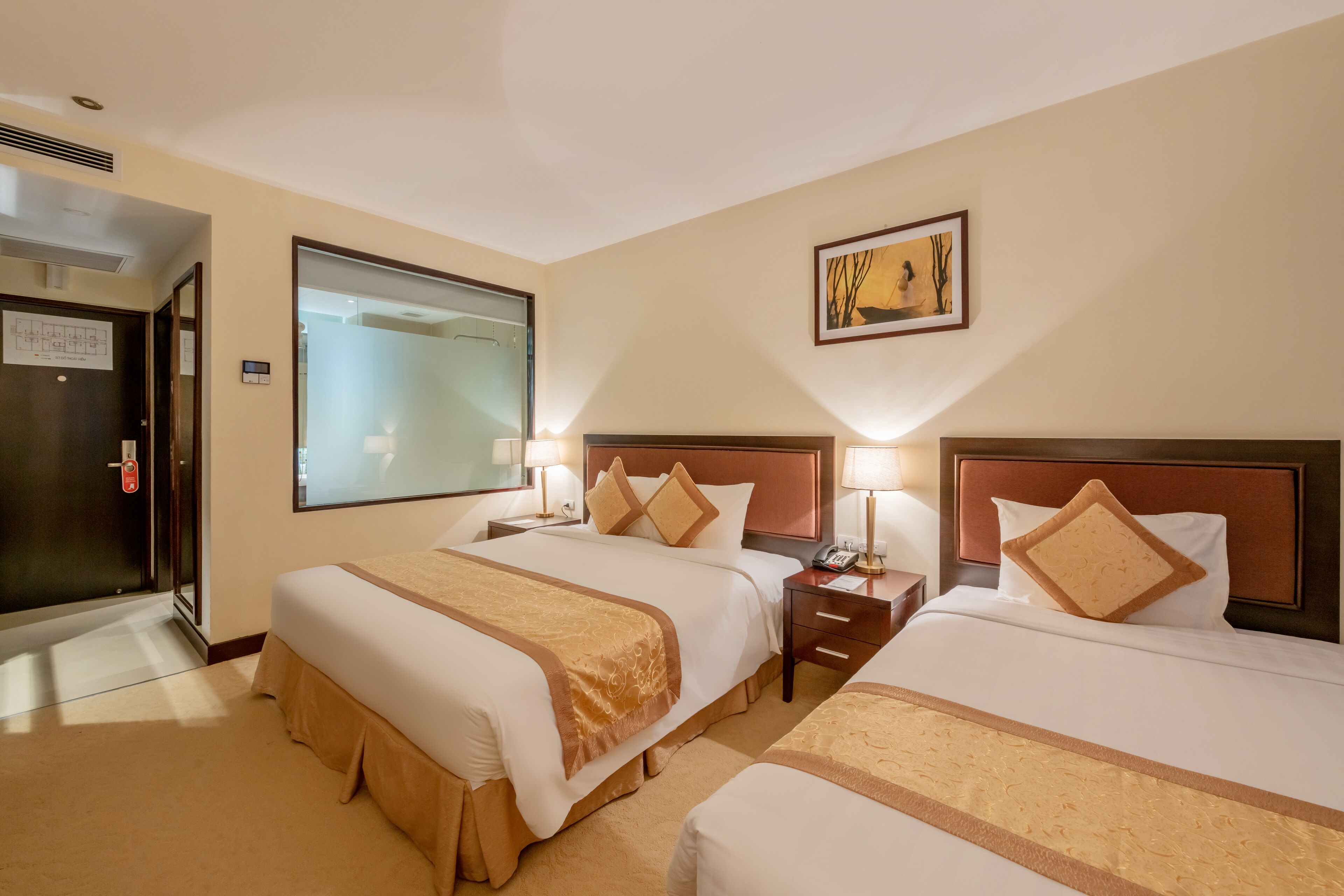 A hotel room with two beds, each with a wooden headboard, bedside tables with lamps, and a large window.