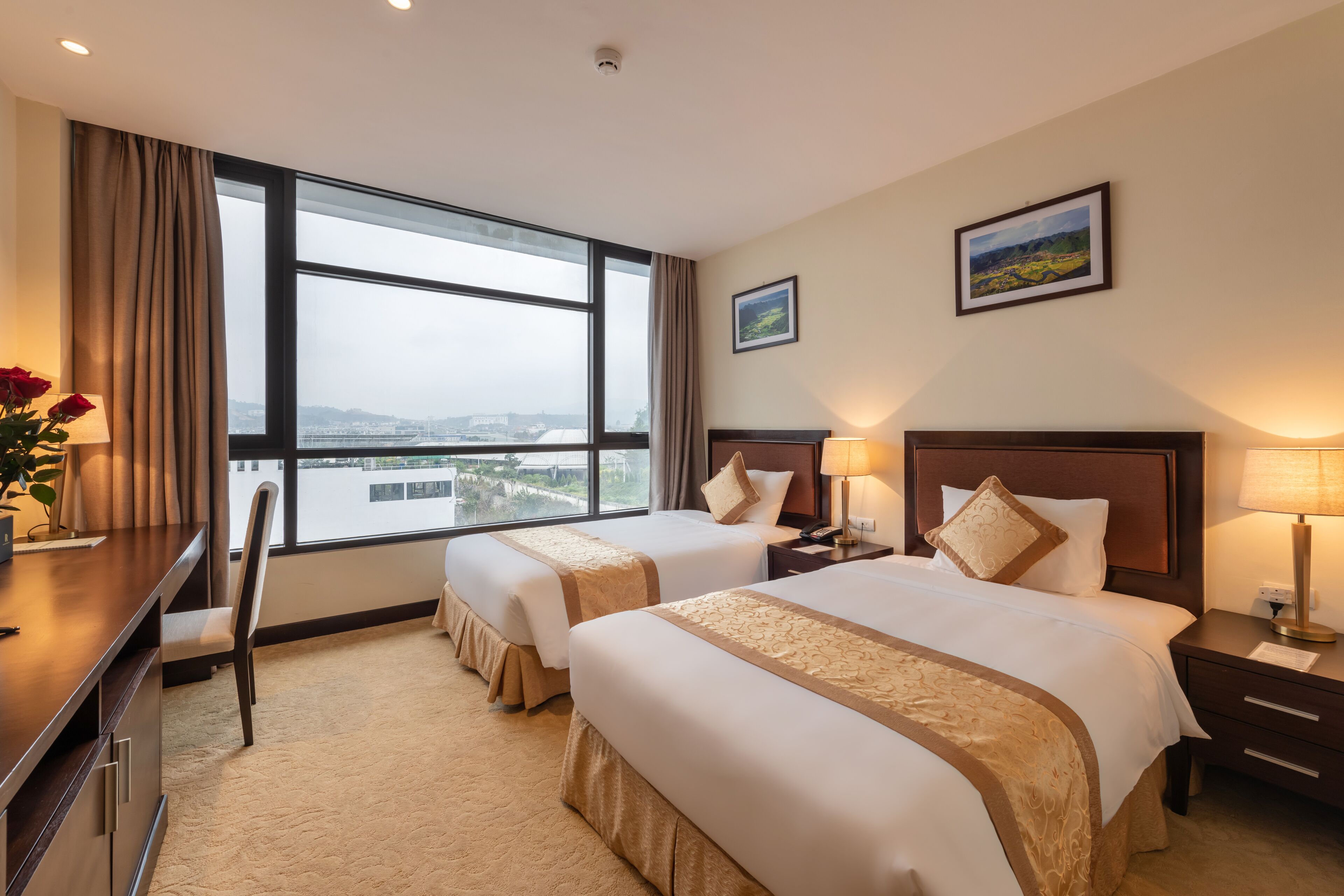 A hotel room with two beds, a desk, a chair, a nightstand, and a large window offering a city view.