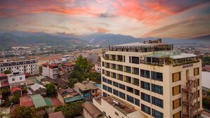 Un hotel con varios pisos, una terraza en la azotea y vistas a un río y montañas al fondo.