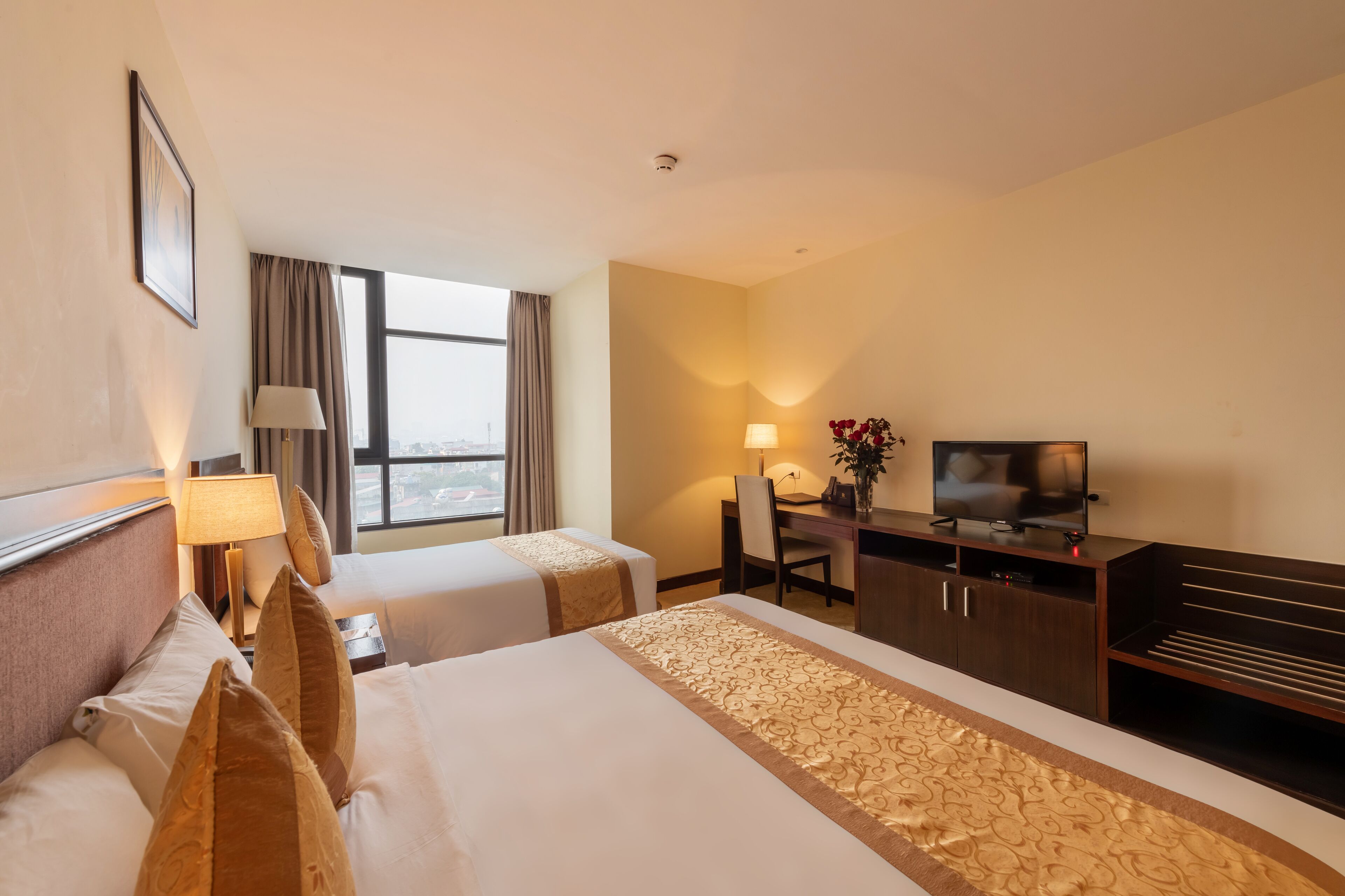 Habitación de hotel con dos camas, un escritorio con televisor y vista a la ciudad a través de amplias ventanas.