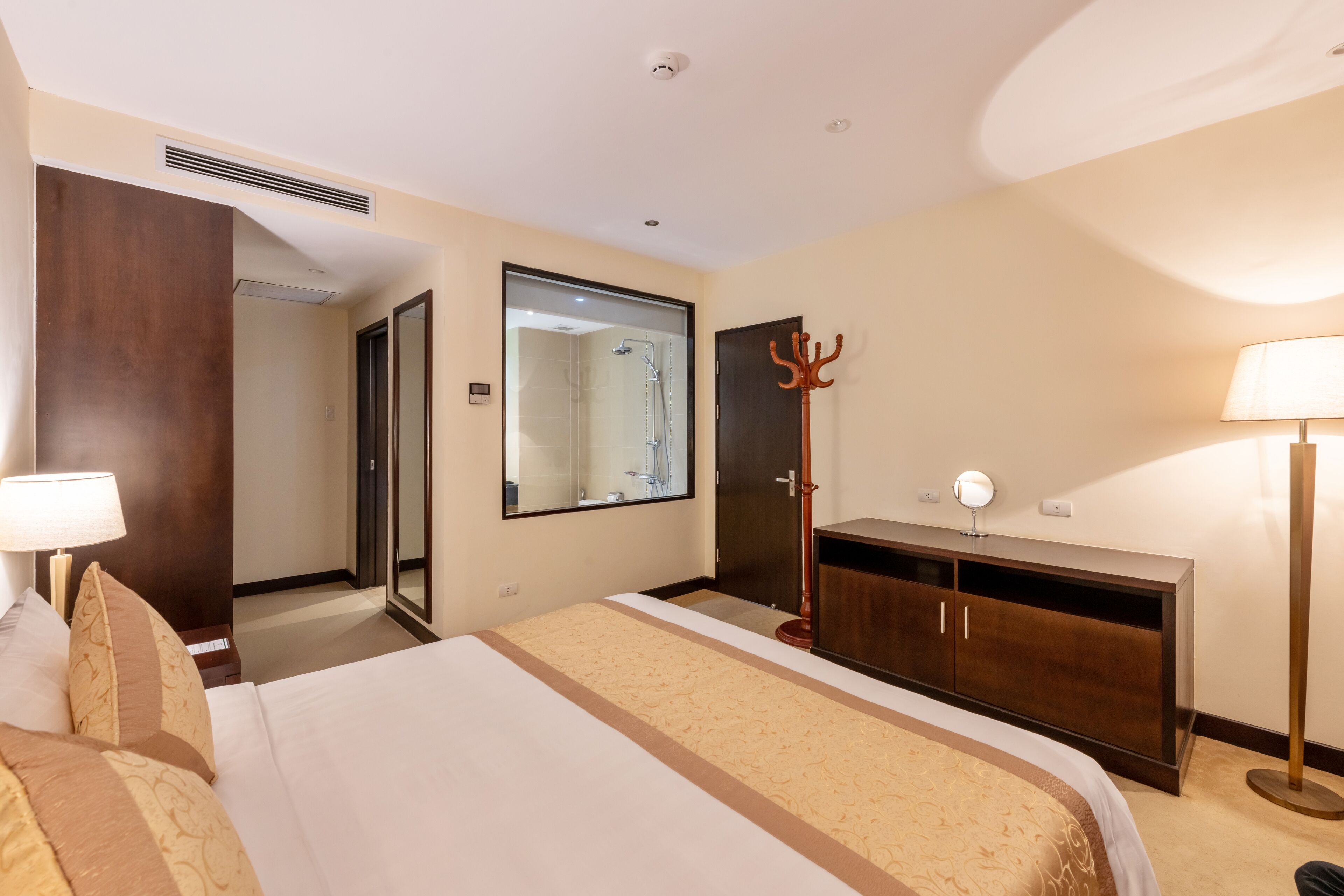 A hotel room with a large bed, a wardrobe, a mirror, a bedside table with a lamp, and a view of the bathroom.
